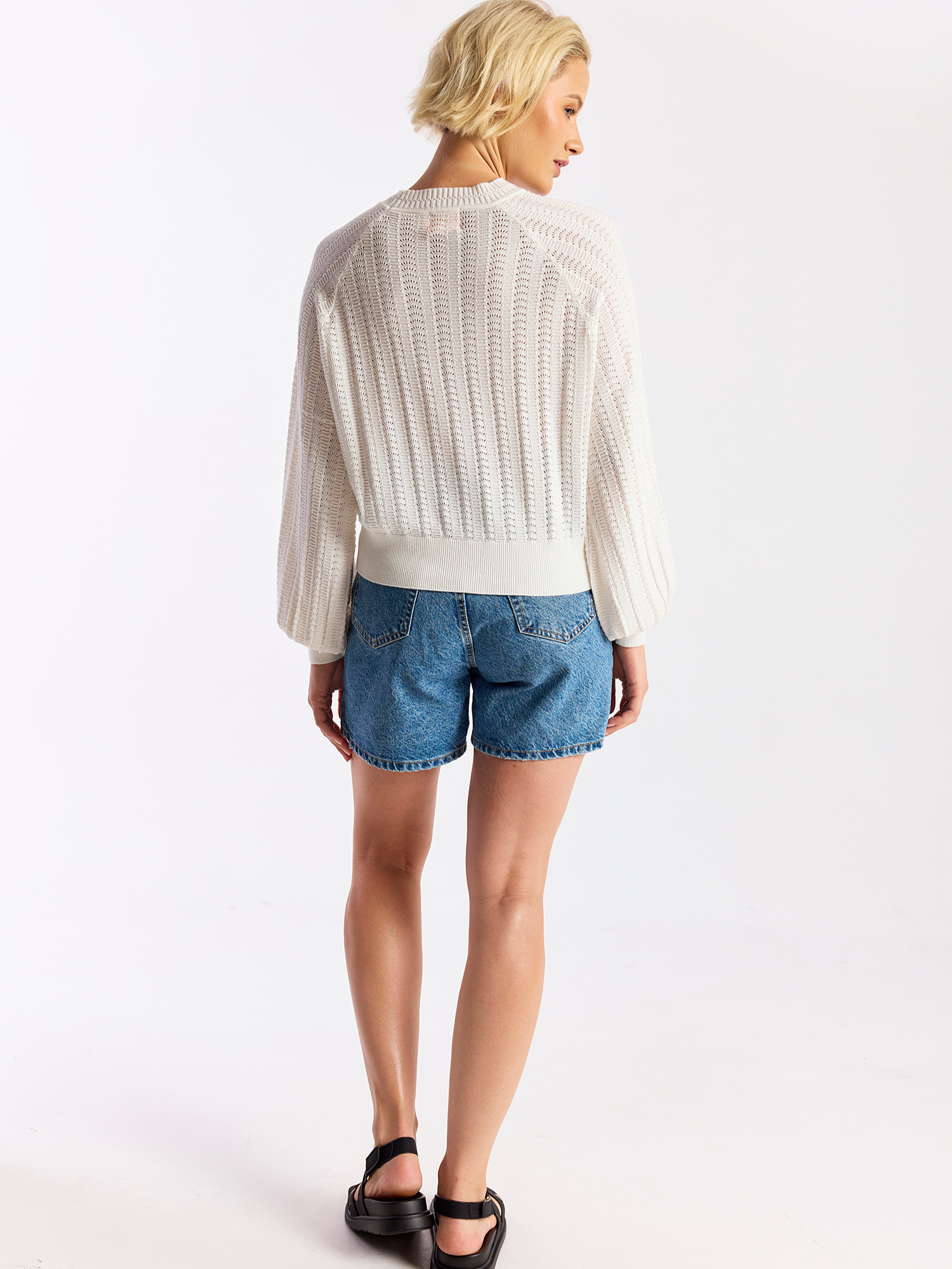 Woman wearing a white lace top and blue denim shorts on a plain background