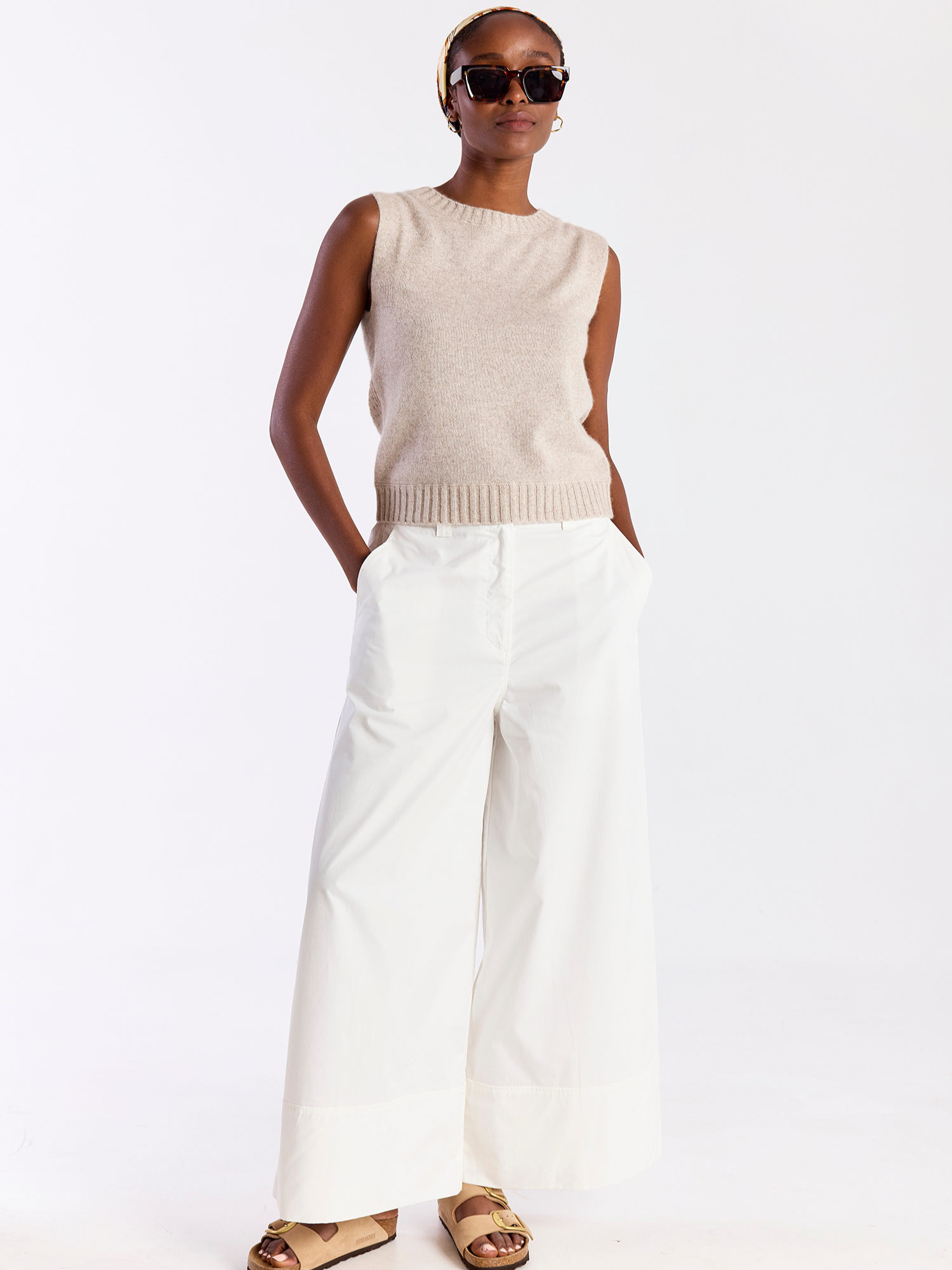 Woman wearing a beige sleeveless top and white pants on a white background