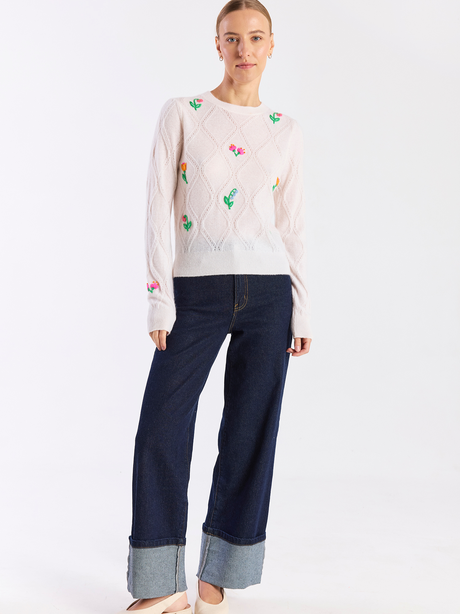 Woman wearing a light pink sweater with floral patterns and dark blue jeans on a white background