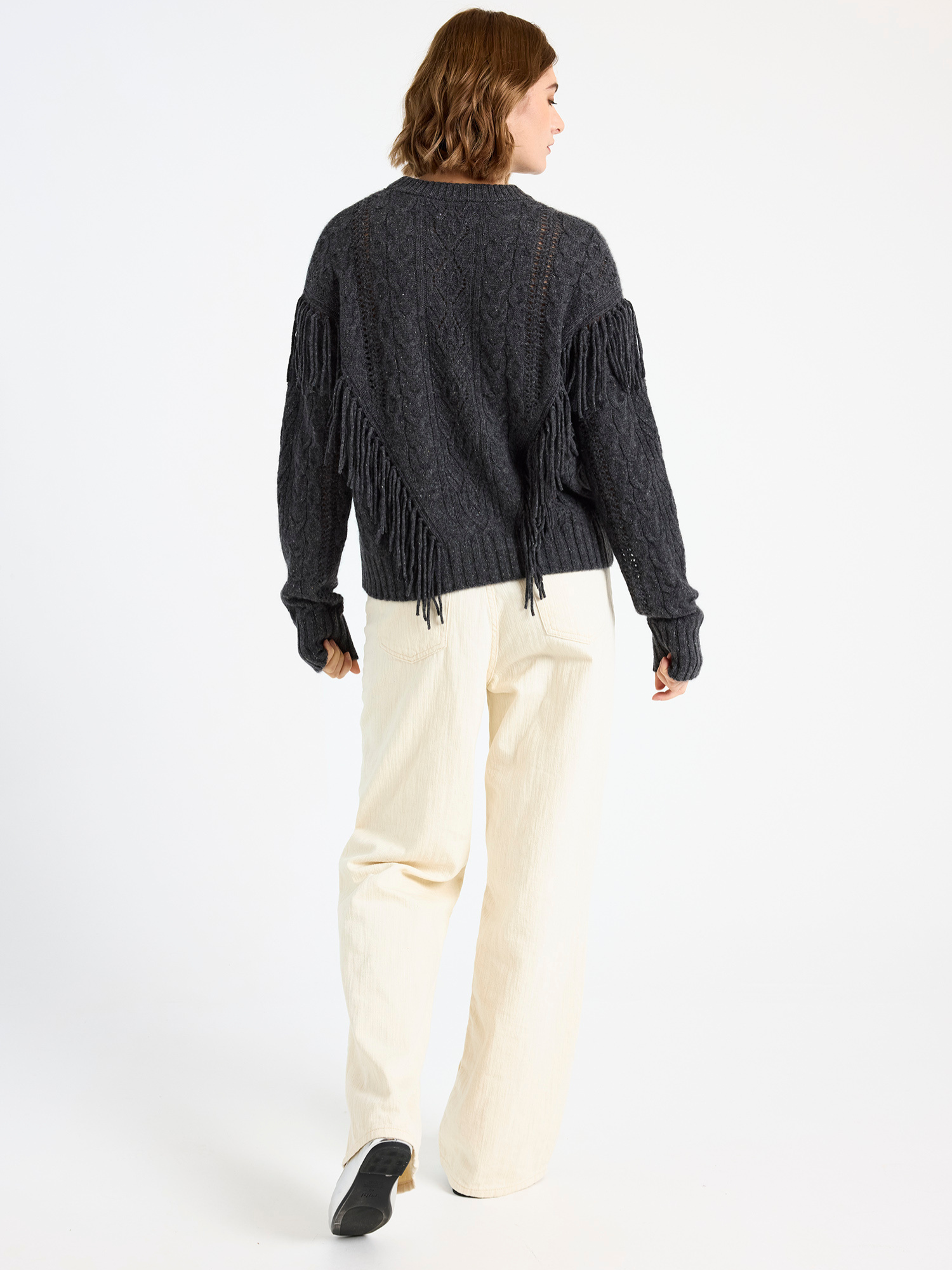 Woman wearing a dark grey cable fringed sweater and ecru jeans on a white background
