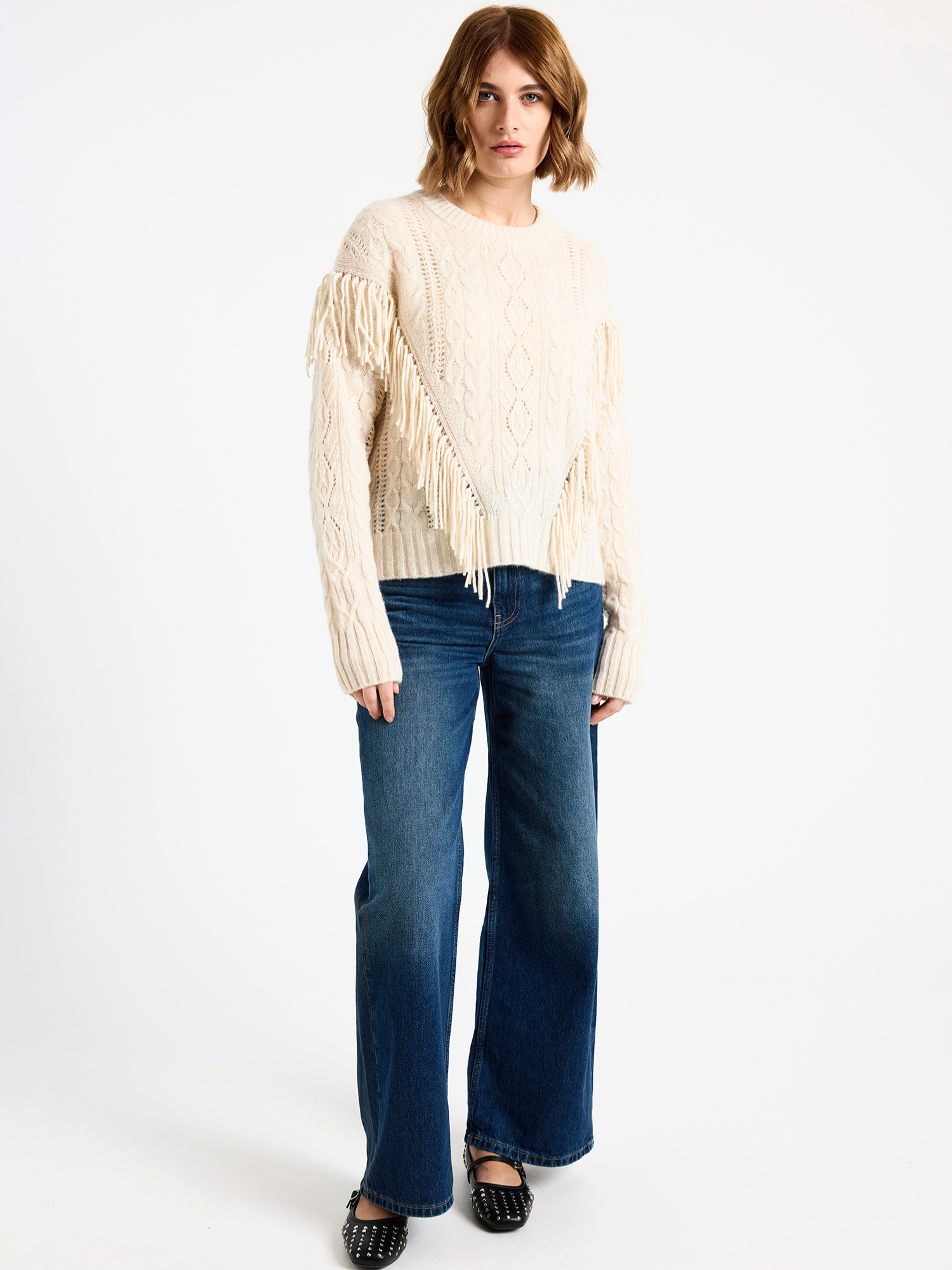 Woman wearing a cream fringed cable sweater and blue jeans on a white background