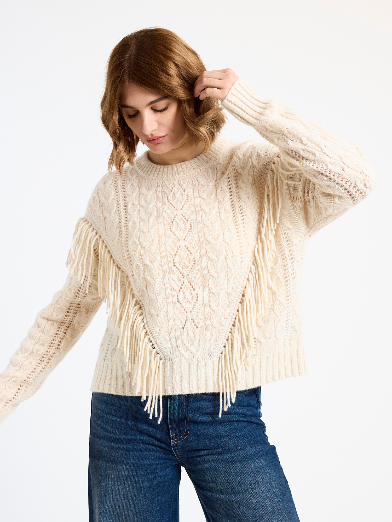 Woman wearing a beige fringe cable sweater and blue jeans on a white background