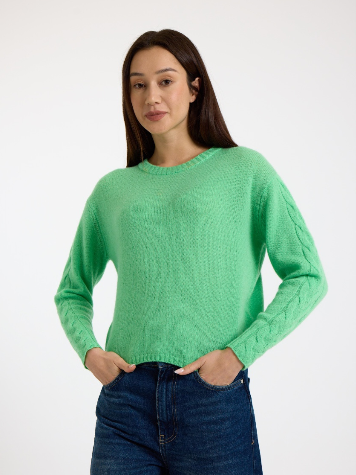 Woman wearing a green sweater with cable detail down the arm and blue jeans on a white background