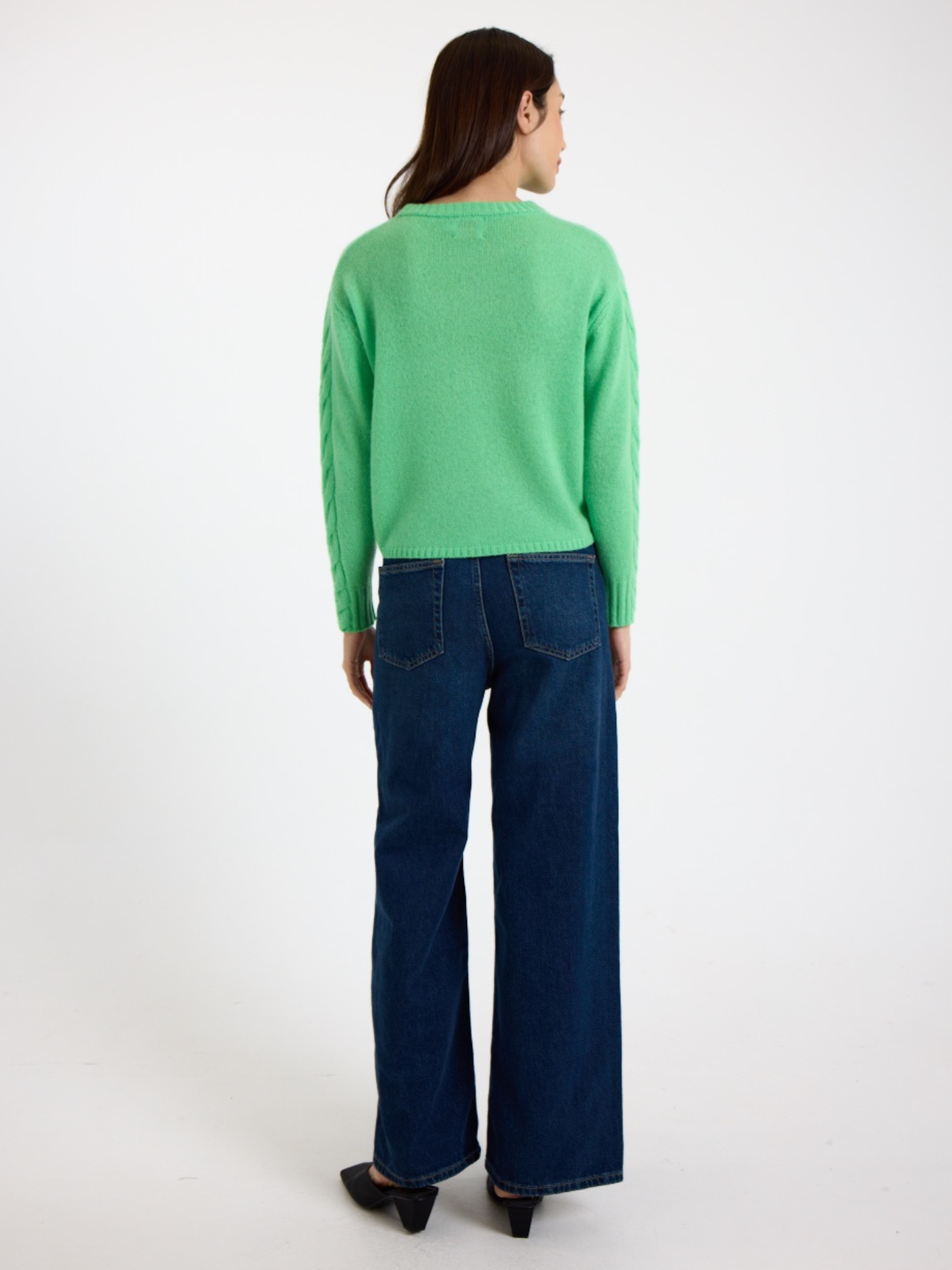 Woman wearing a green sweater with cable detail down the arm and blue jeans on a white background