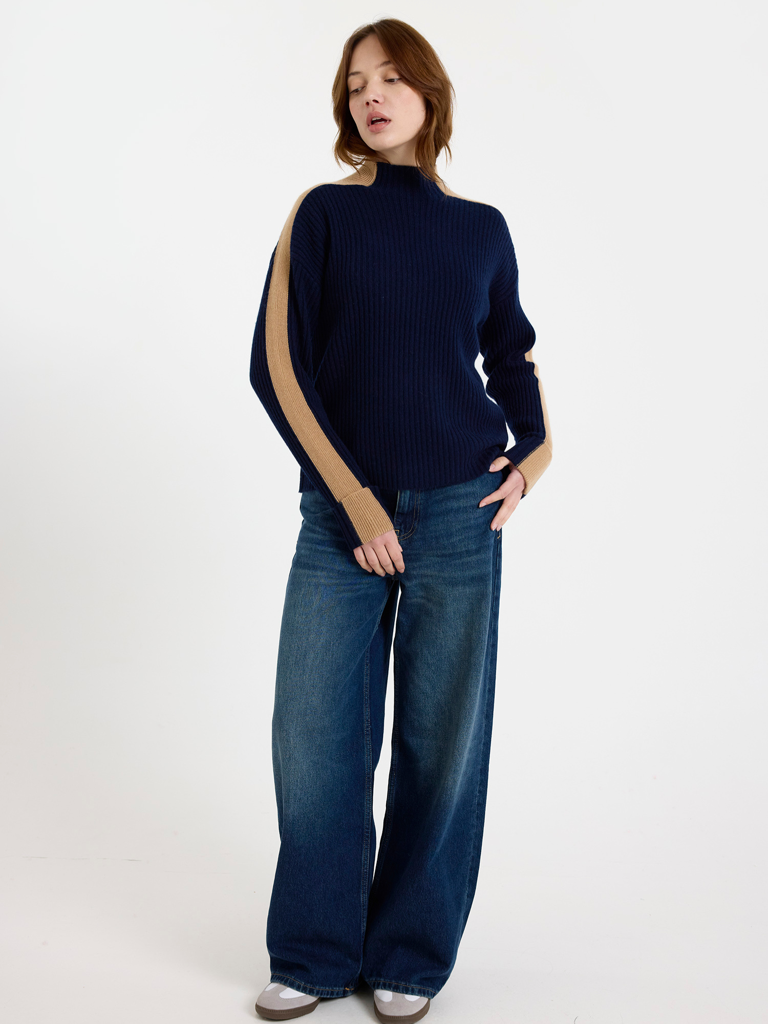 Woman wearing a navy ribbed sweater with camel colour stripe down the arm and blue jeans on a white background