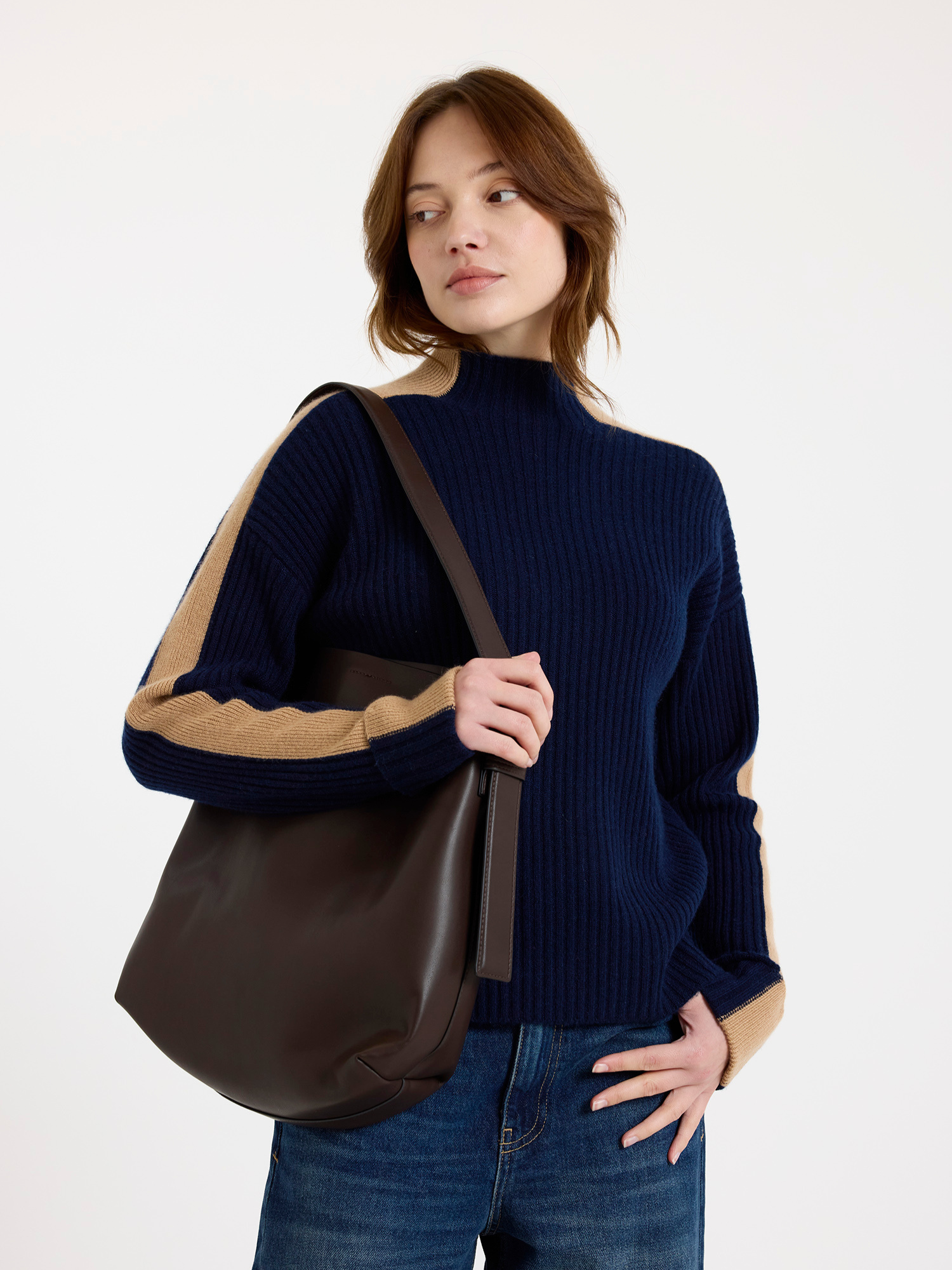 Woman holding a brown leather bag wearing a navy ribbed mock neck sweater with camel colour stripe down the arm against a white background