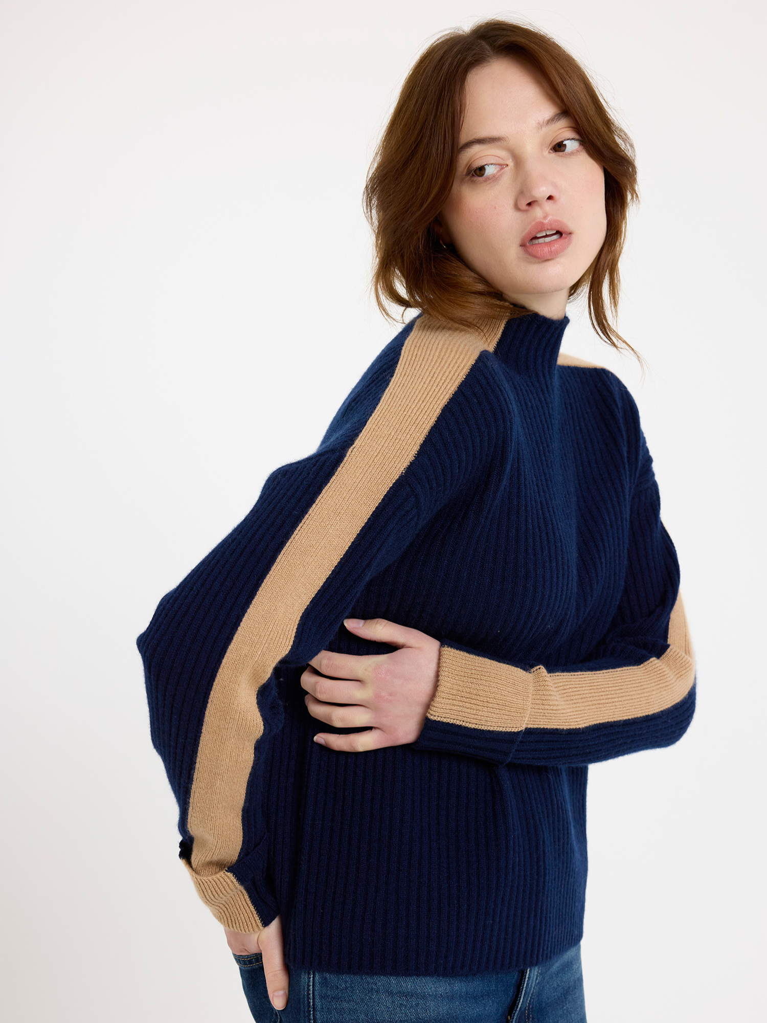 Woman wearing a navy ribbed sweater with camel colour stripe down the arm and blue jeans on a white background