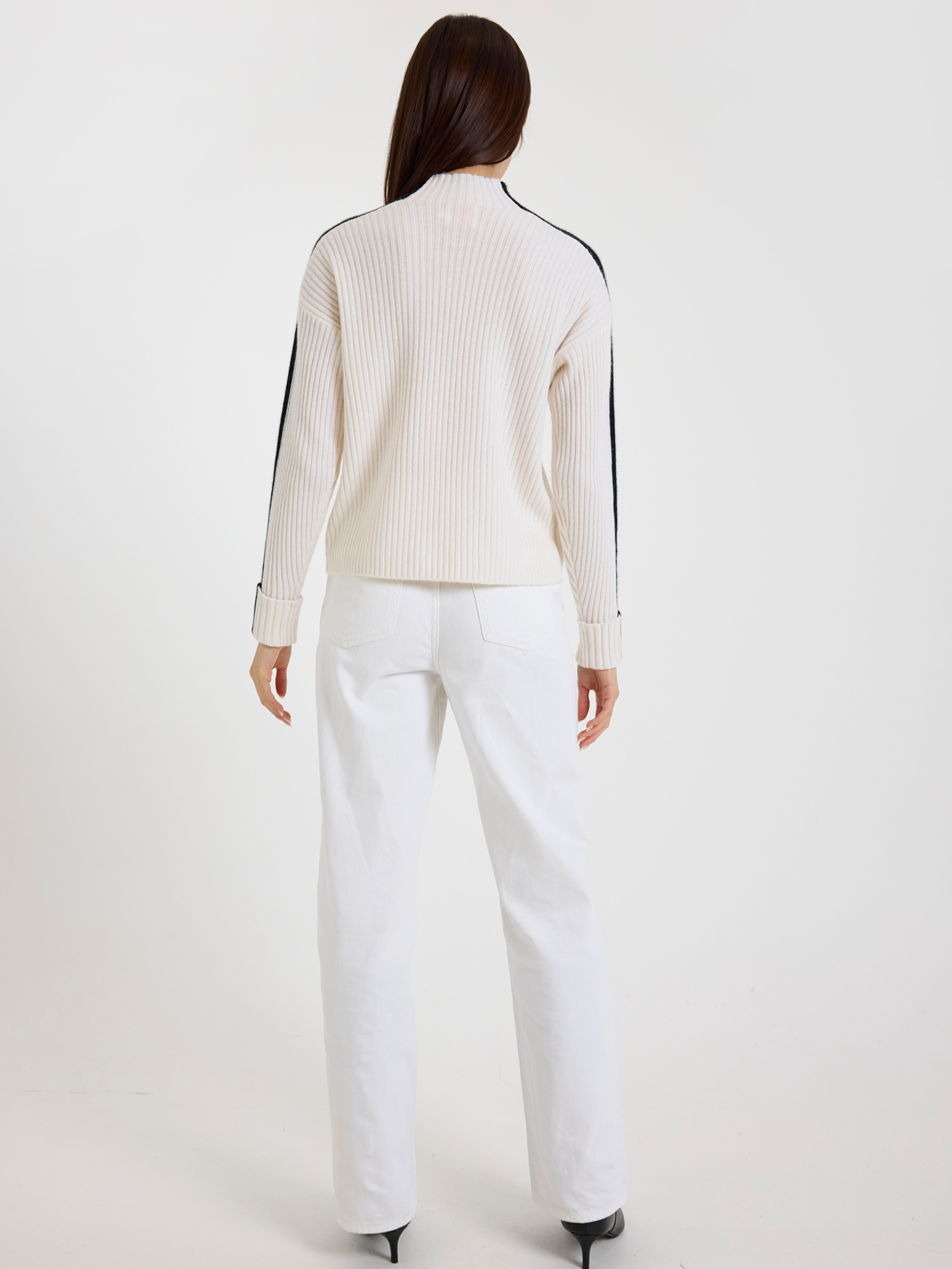 Woman wearing a white ribbed sweater with black stripe sleeves and white jeans pants on a white background