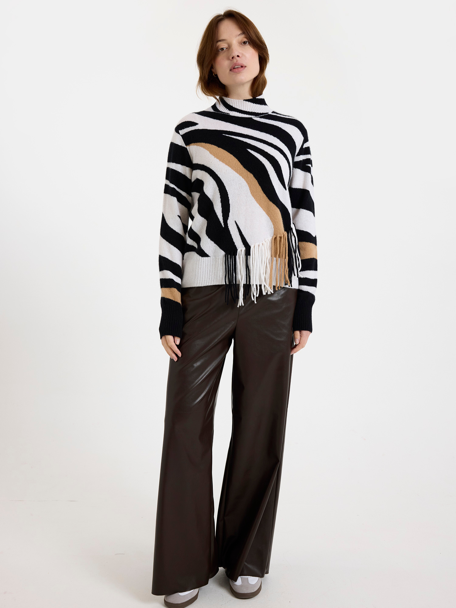 Woman wearing a black and white zebra pattern sweater with fringes and brown leather trousers on a white background
