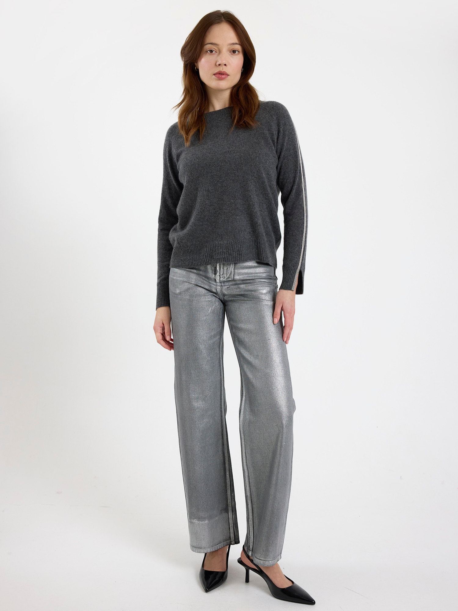 Woman wearing a grey sweater with glitz detail down the arm and silver jeans on a white background