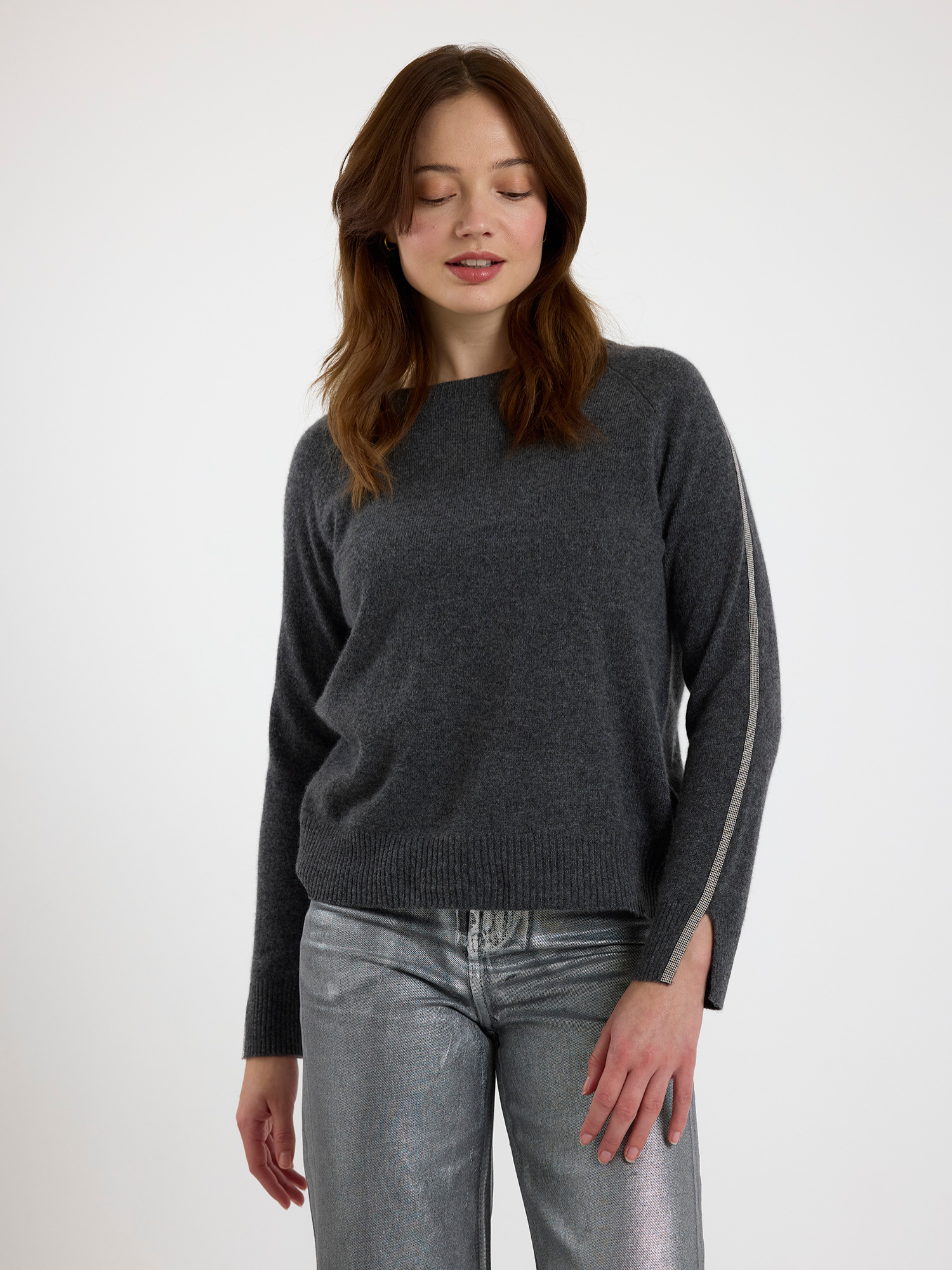 Woman wearing a grey sweater with glitz detail down the arm and silver jeans on a white background