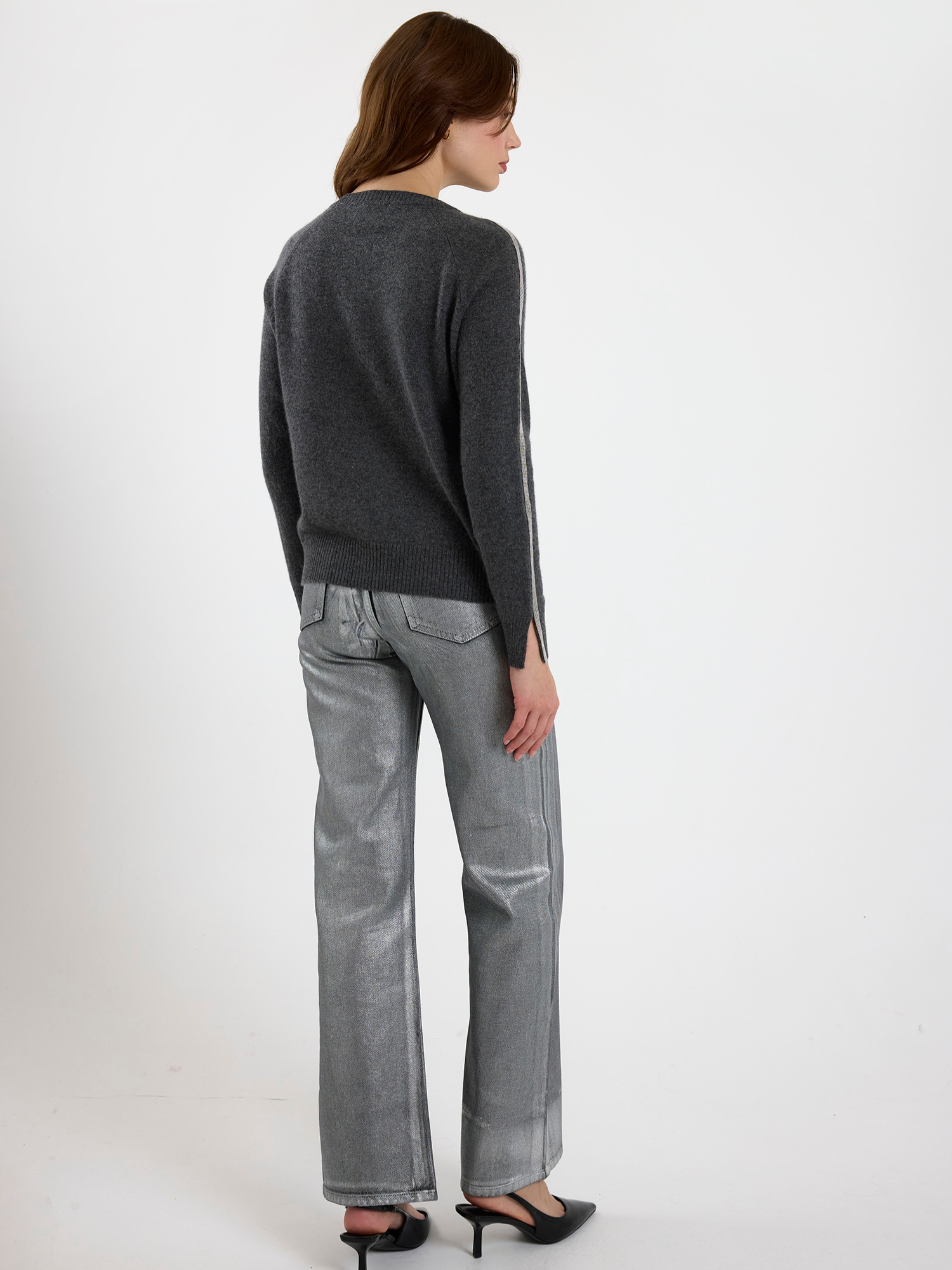 Woman wearing a grey sweater with glitz detail down the arm and silver jeans on a white background