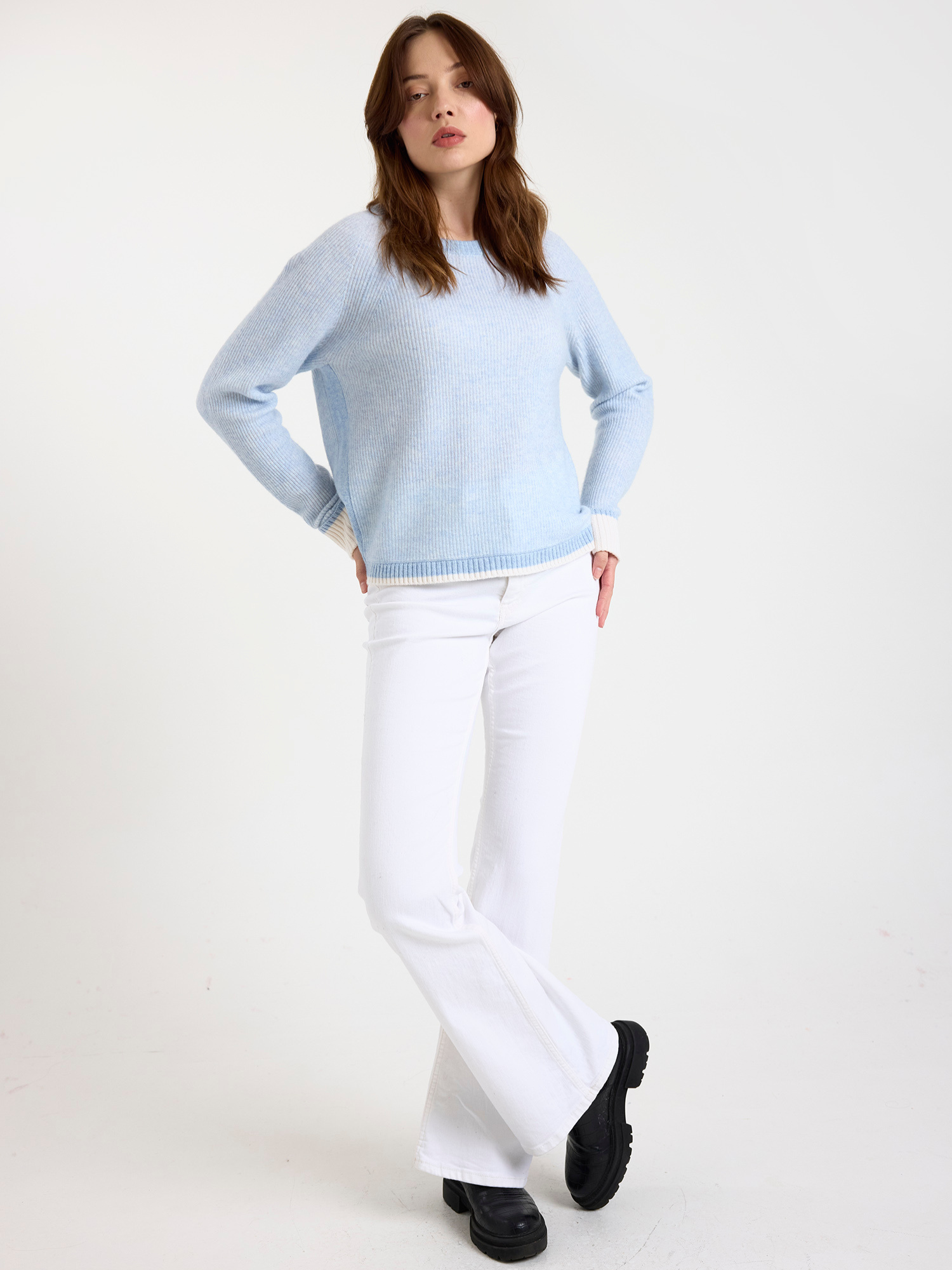Woman wearing a ribbed light blue sweater and white jeans on a white background