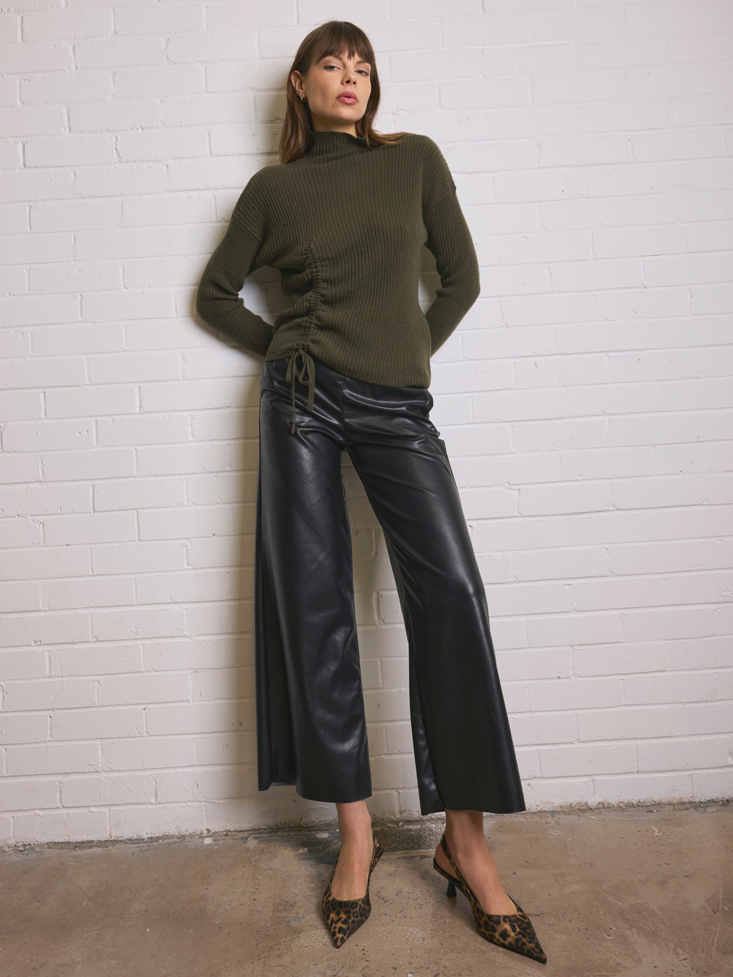 Woman wearing a green sweater and black leather pants against a white brick wall.