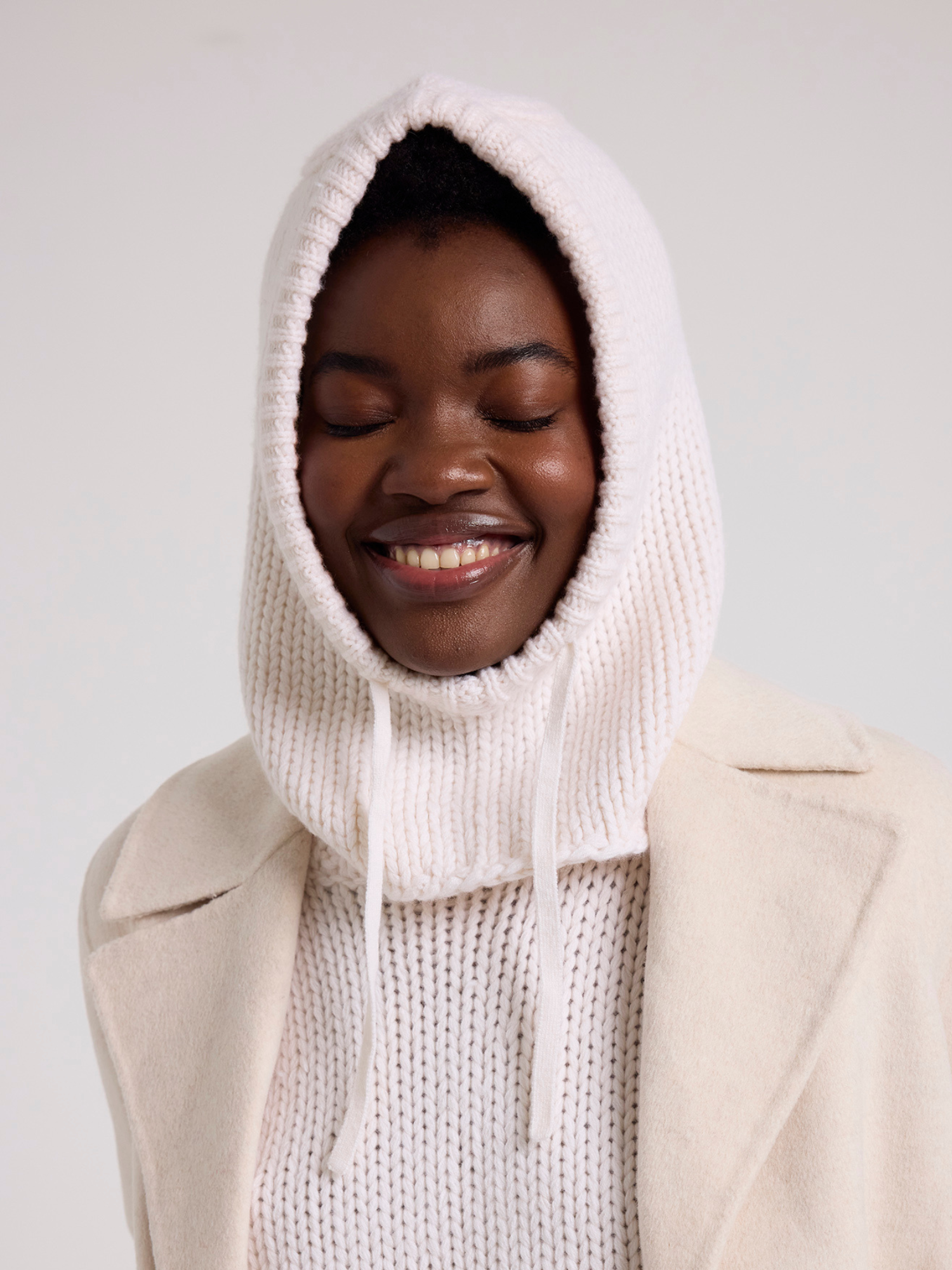 Person wearing a white knitted balaclava over a beige coat on a plain background
