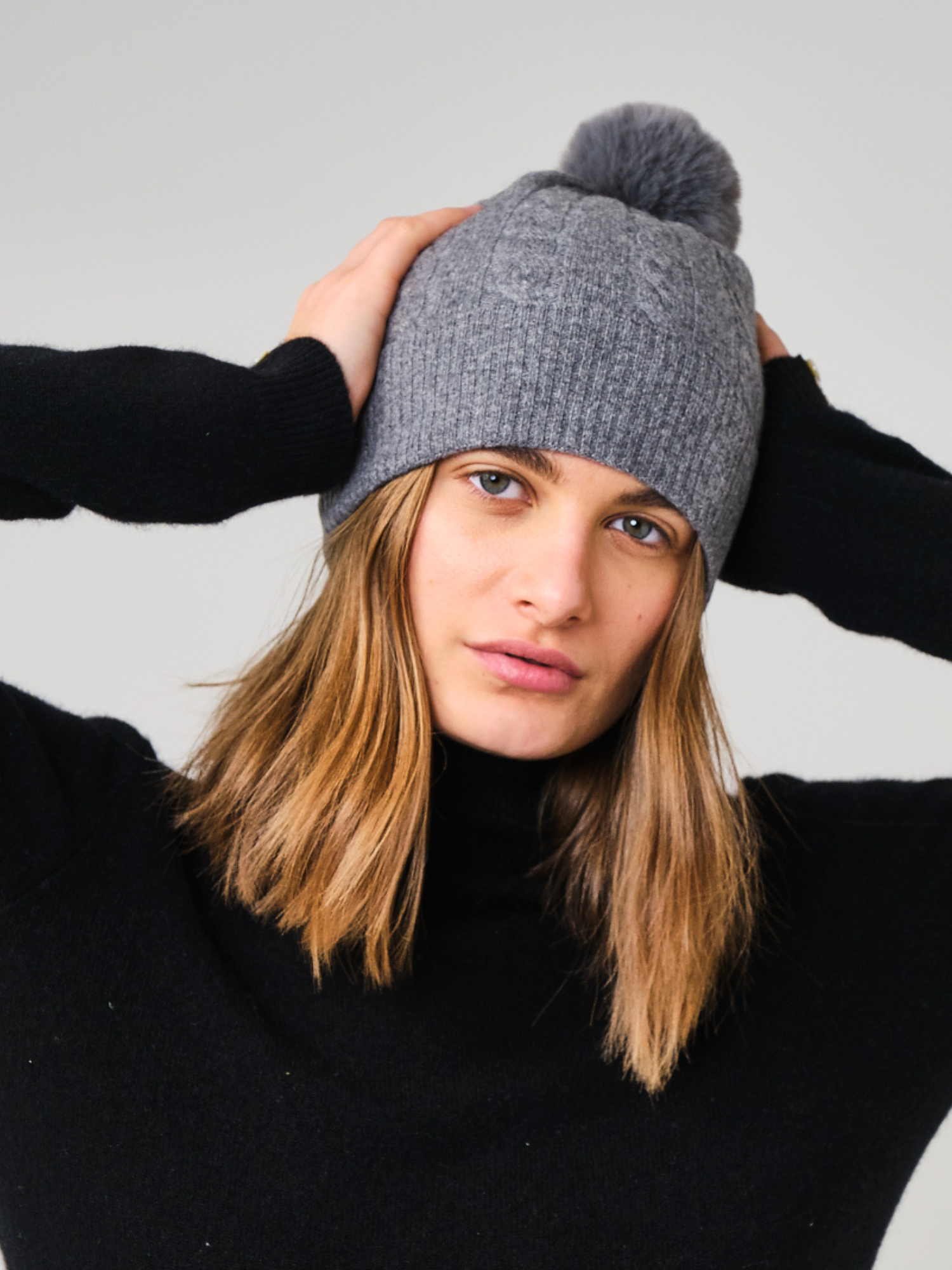 Person wearing a grey knit beanie with a pom-pom on a plain background