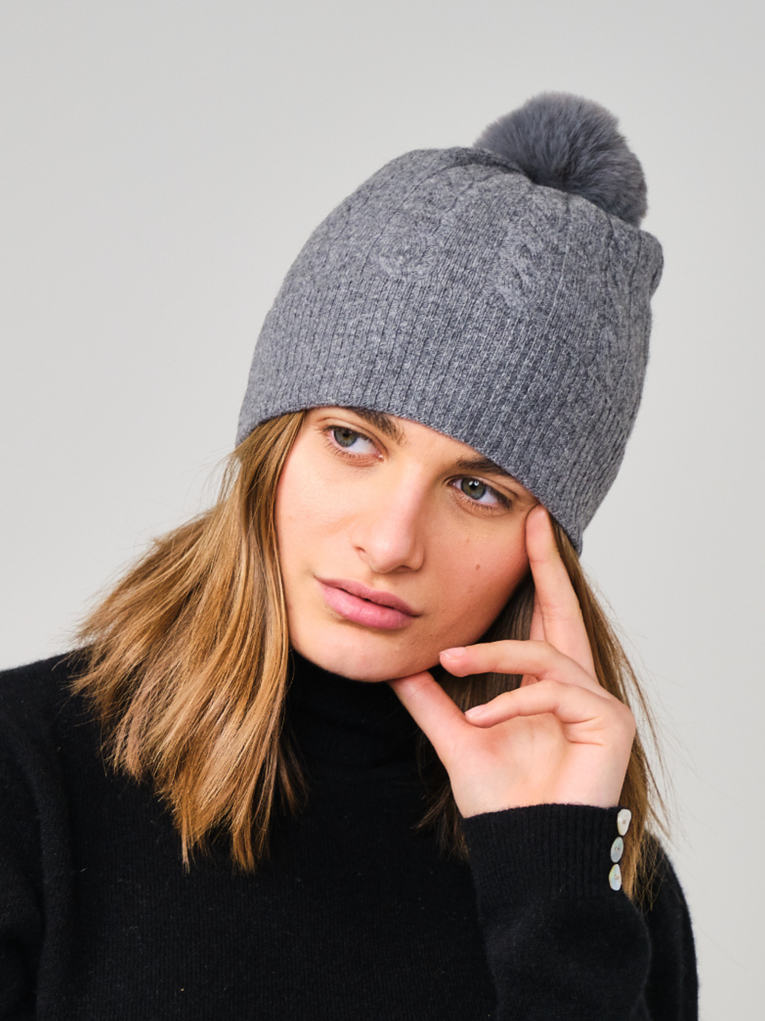 Person wearing a grey knit beanie with a pom-pom on a plain background